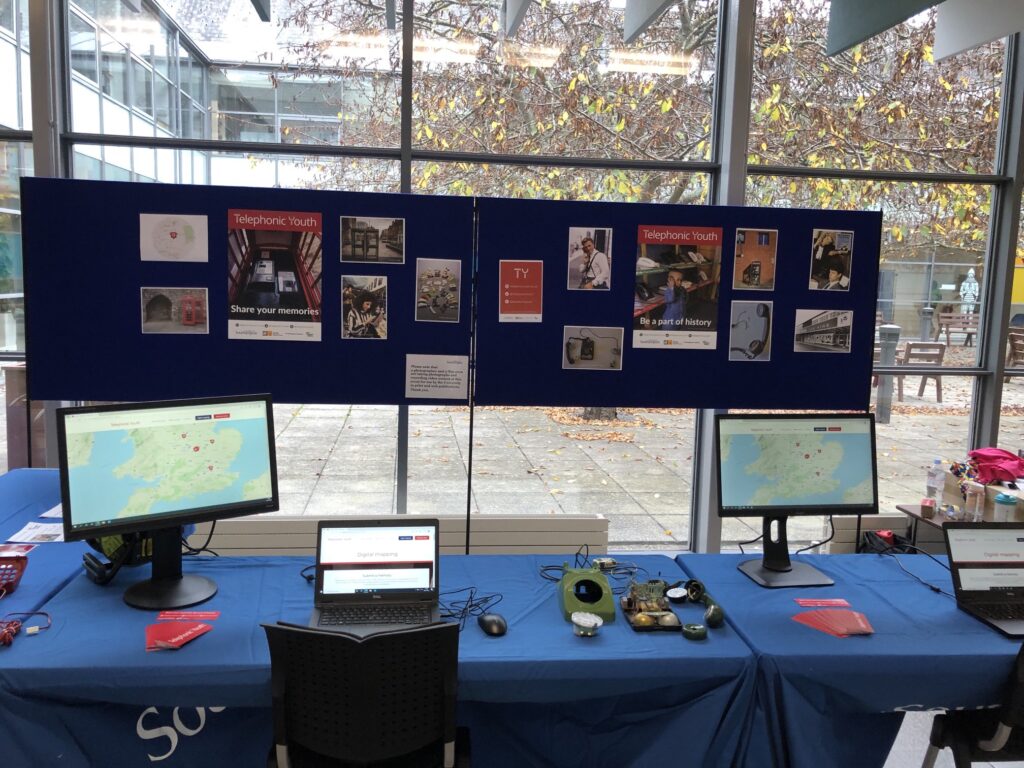 Image shows three tables in an 'L' formation with blue Southampton university-branded table cloths. On top of the tables are two laptops, two computer screens, and three disassembled phones. Behind the tables are two large poster boards with a number of posters featuring images of telephone users from the 1980s and 1990s. The stall is in front of a set of windows which look out onto a courtyard at the University of Southampton.