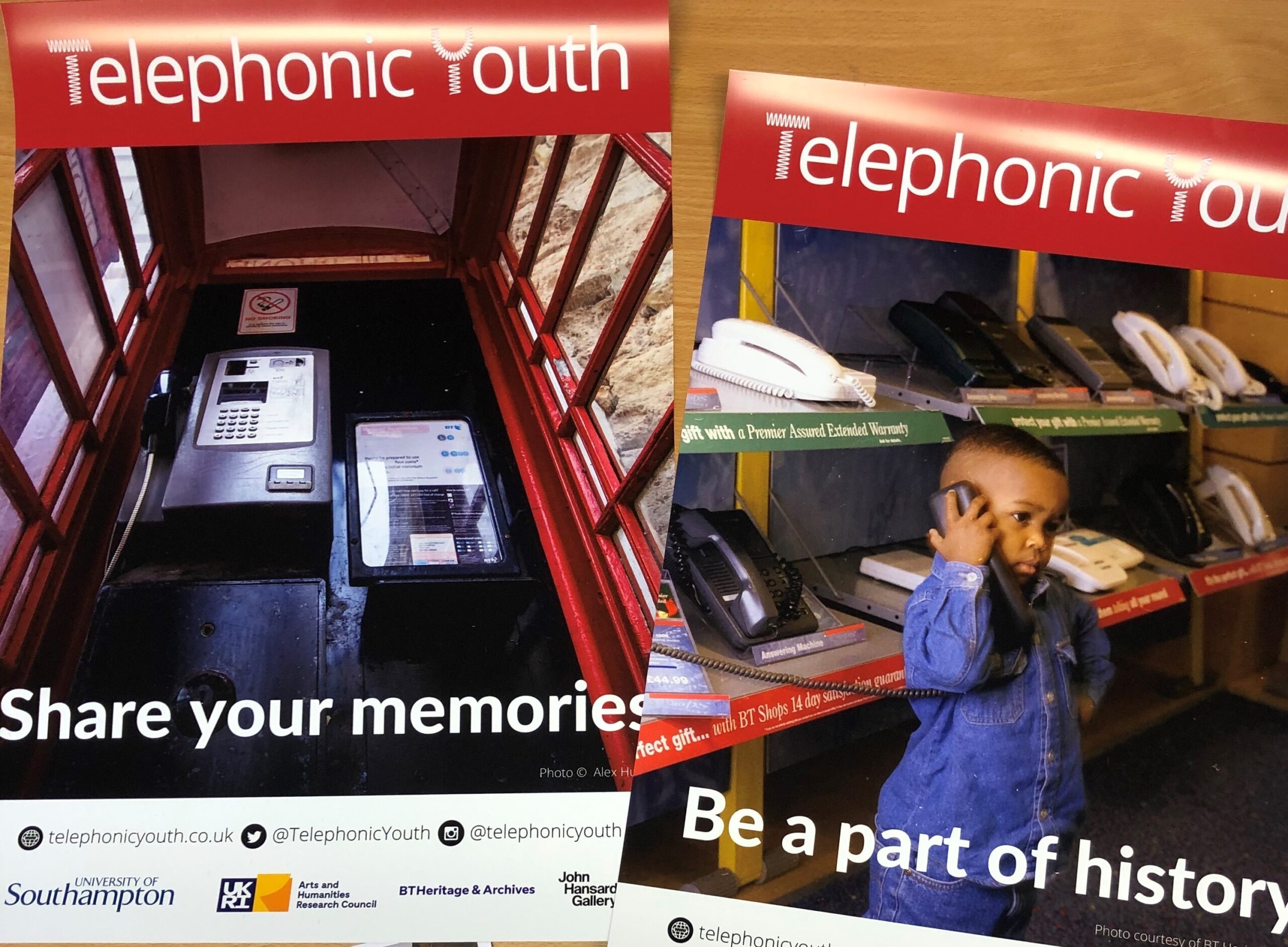 Image shows Telephonic Youth branded posters laid out on a table. The posters include the phrases 'Share your memories' and 'Be a part of history'. One has an image of the interior of a red telephone box; the second has an image of a young boy playing with a telephone in a shop.