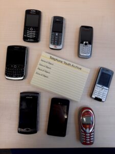 A set of 8 old mobile phones laid out in a circle on a light wooden table. In the middle are a set of 'Telephonic Youth Archive' tags which ask for the name, date, place, and description of an object