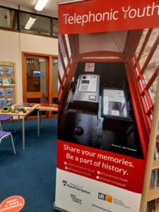 Image shows a pop-up banner. pull-up banner featuring a large image of the interior of a red phone box, the logo for the Telephonic Youth project and the text 'Share your memories. Be a part of history'. Also included on the banner are logos for the project's funders and partners, and contact details (@telephonicyouth on Twitter, Facebook, and Instagram; and telephonicyouth.co.uk). Behind the banner is a set of tables in the library, a blue carpet and light coloured walls.