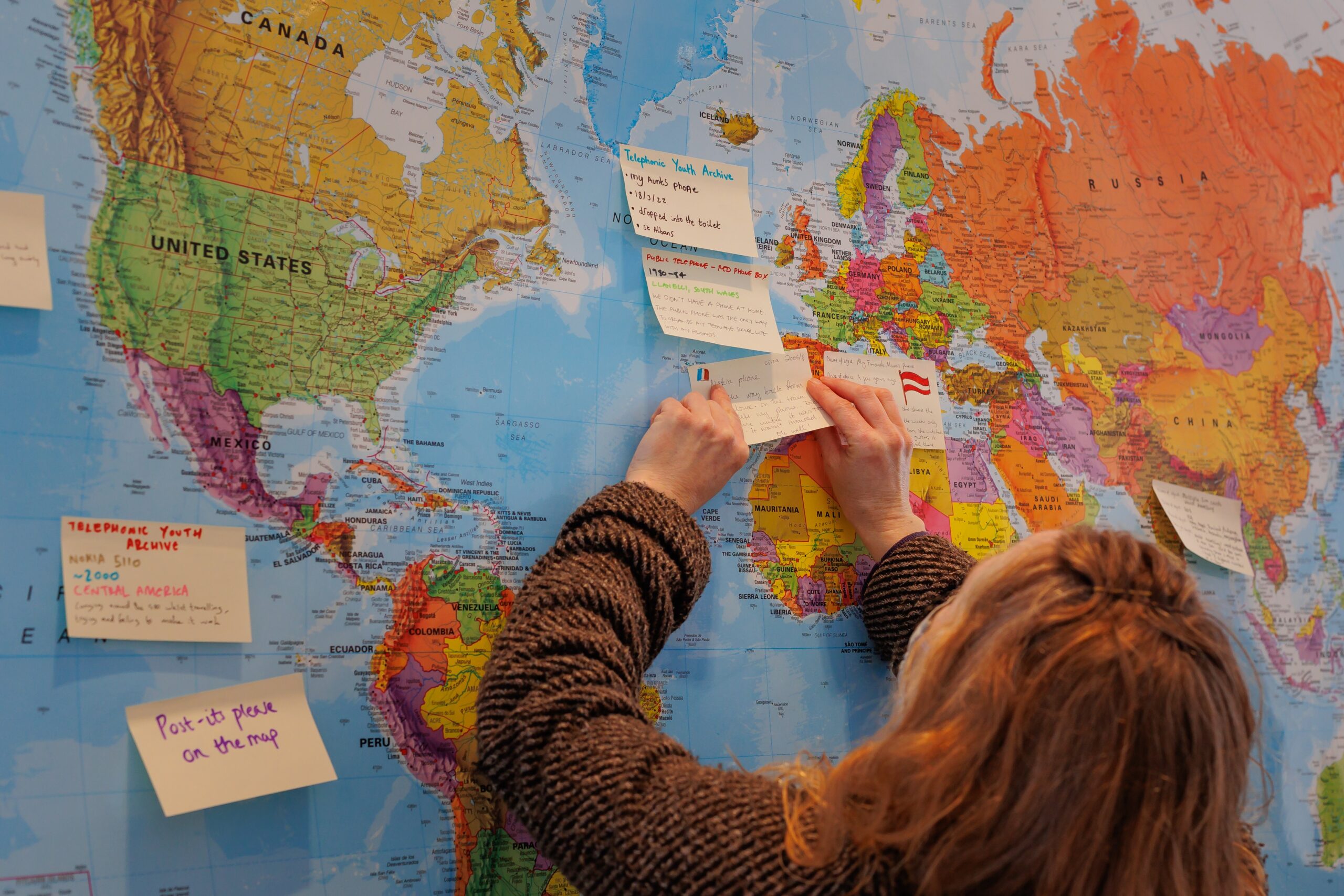 A white woman with medium length, red-ish hair and wearing a brown coat and blue disposable face mask is putting a post-it note on a map of the world. There are a number of post-its on the map, each with a memory relating to a telephone. The post-its have 'Telephonic Youth Archive' written at the top. The woman's memory appears to be related to a place in France and has a small French flag drawn in pen in the corner.