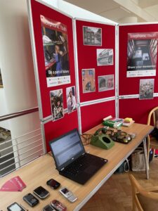 Image shows a table with several old mobile phones, a disassembled green rotary phone, and a laptop along with other materials (including post-it’s and bookmarks). Behind the table is a set of poster boards with Telephonic Youth branded posters and images of 1980s and 1990s telephone users from the BT archives and images of Southampton.