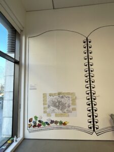 The A1 map and memories stuck onto a wall with a large outline in the form of a wirebound book painted on it. Other small artworks are also included in the book, including some painted leaf shapes. To the left is a large window looking out onto a public open space.