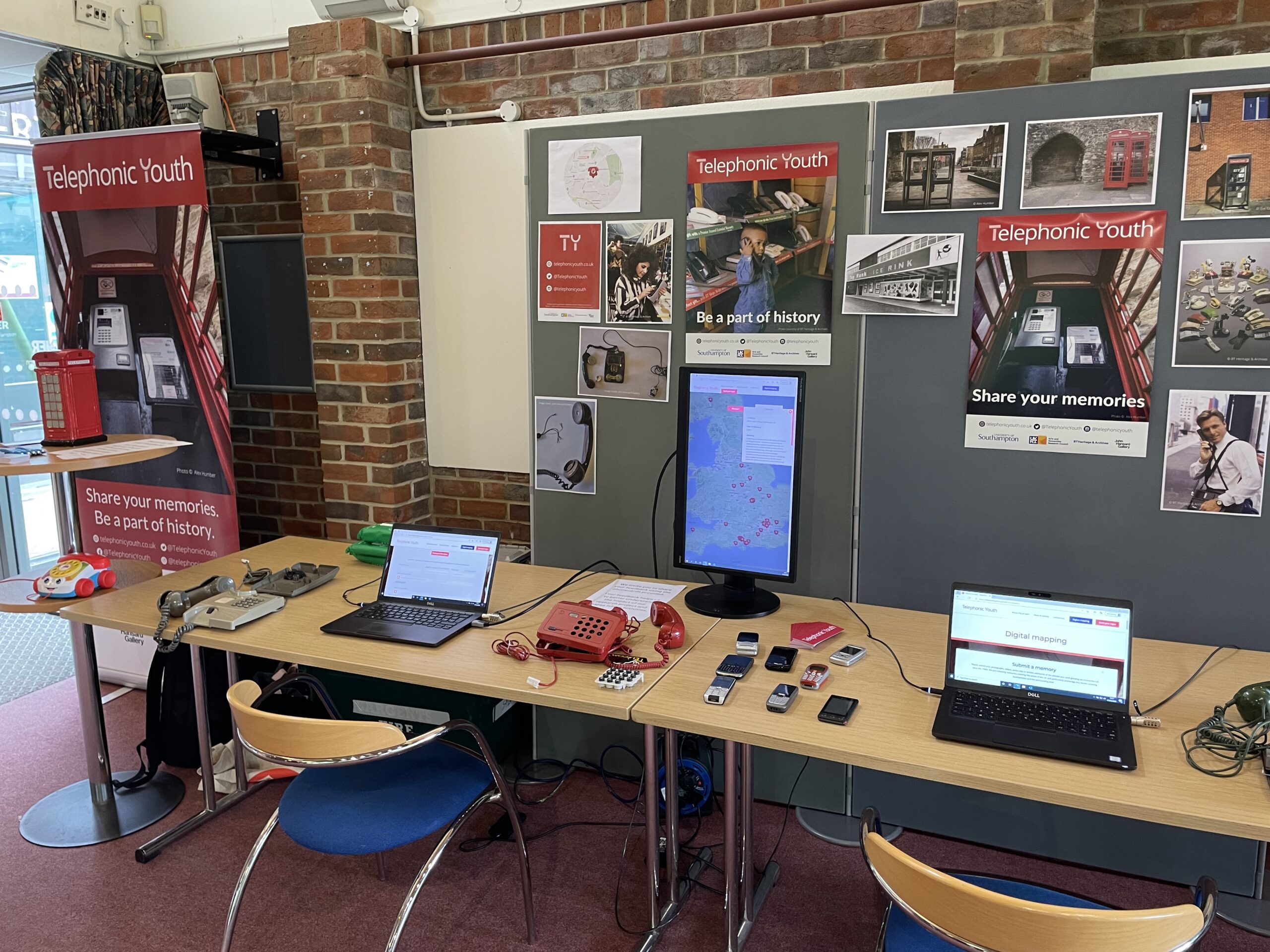 Two laptops, three disassembled landline telephones, and eight mobile phones laid out on two tables. In the background are various posters from BT or places Southampton