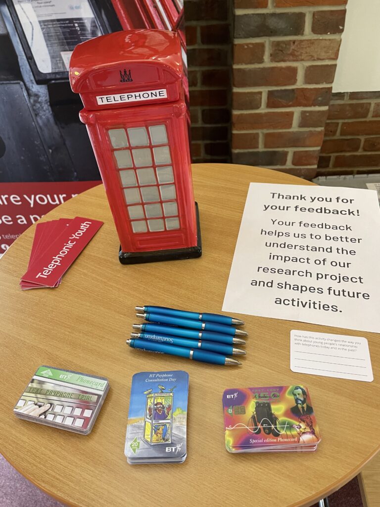 A red phone box-shaped cookie jar and set of replica phonecards on a circular table with a sign requesting feedback